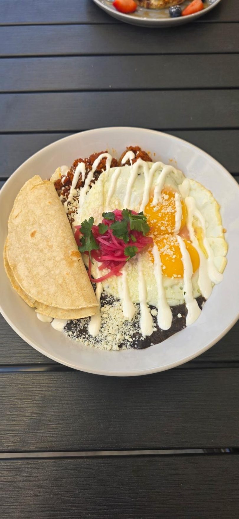A white plate of chilaquiles with fried eggs, black beans, tortillas, and pickled onions.